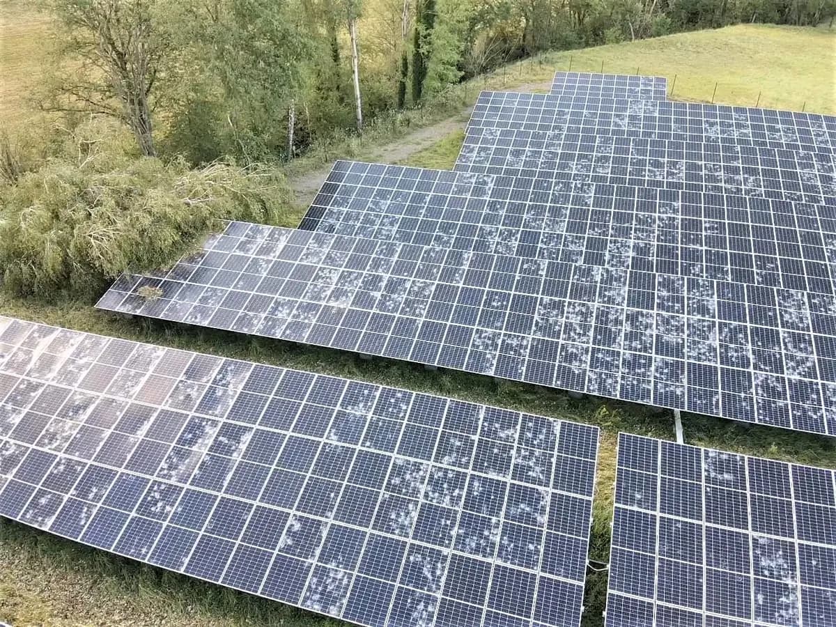 Vue aérienne d'un parc photovoltaïque au sol dont les modules portent de nombreux impacts de grêle, visibles en marques claires sur les cellules.